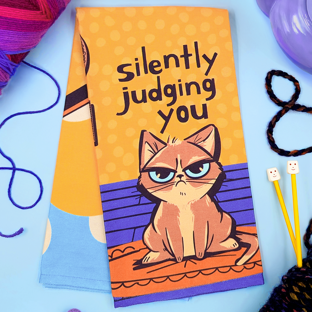 Tea towel with a cat illustration and text 'silently judging you' on a blue background with yarn and knitting tools.