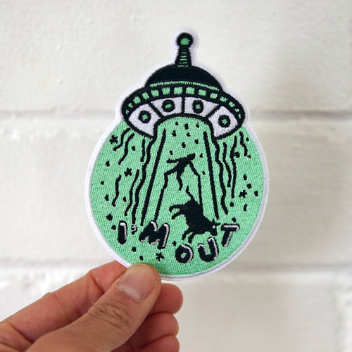 Hand holding a green and black patch with alien design and text.