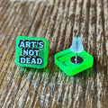 Green badge with 'Art's Not Dead' text on a wooden surface, featuring the brand 'Rapscallion'.